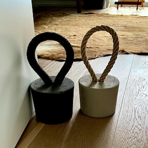 Può includere: Due fermaporta cilindrici. Uno è nero con un manico in corda nera, l'altro è grigio chiaro con un manico in corda beige. Entrambi i fermaporta sono su un pavimento in legno.
