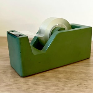May include: A matte sage green desktop tape dispenser with a rectangular base and a central circular hub holding a roll of clear adhesive tape. The dispenser features a metal serrated cutting edge at the front for tearing the tape.