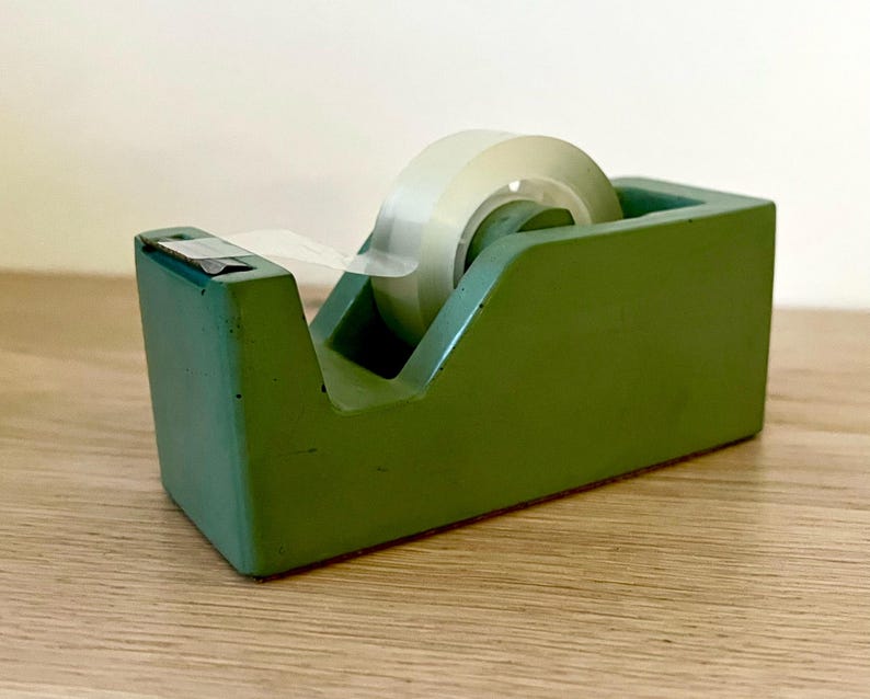 May include: A matte sage green desktop tape dispenser with a rectangular base and a central circular hub holding a roll of clear adhesive tape. The dispenser features a metal serrated cutting edge at the front for tearing the tape.