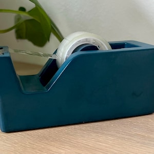 May include: A dark teal desktop tape dispenser with a weighted base, featuring a clear plastic serrated cutting edge at the front and a roll of transparent adhesive tape loaded onto the central spindle, ready for office or craft use.