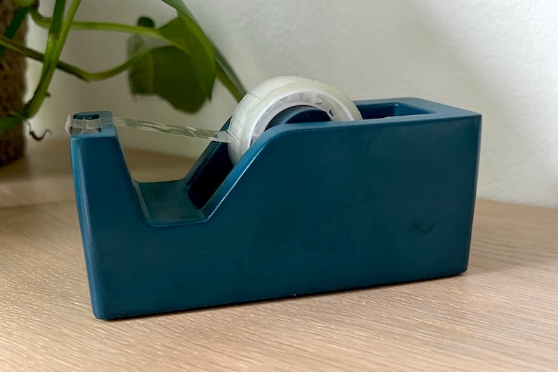 May include: A dark teal desktop tape dispenser with a weighted base, featuring a clear plastic serrated cutting edge at the front and a roll of transparent adhesive tape loaded onto the central spindle, ready for office or craft use.