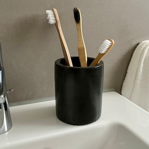 Może przedstawiać: Czarny ceramiczny stojak na szczoteczki do zębów z trzema szczoteczkami do zębów w środku. Dwie szczoteczki do zębów mają białe włosie, a jedna ma czarne włosie.