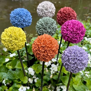 Könnte beinhalten: Dekorative Gartenstecker mit strukturierten, kugelförmigen Blüten in verschiedenen Farben: Blau, Grau, Rot, Pink, Lila, Gelb, Grün und Orange. Die Stecker sind in einem Beet aus grünem Laub und weißen Blumen platziert, mit einem Teich im Hintergrund.