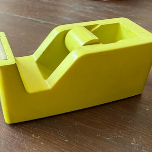 May include: A lime green tape dispenser featuring a smooth, solid-colored plastic body with a built-in metal serrated cutting blade at the front and a central cylindrical core designed to hold a standard roll of adhesive tape for office use.
