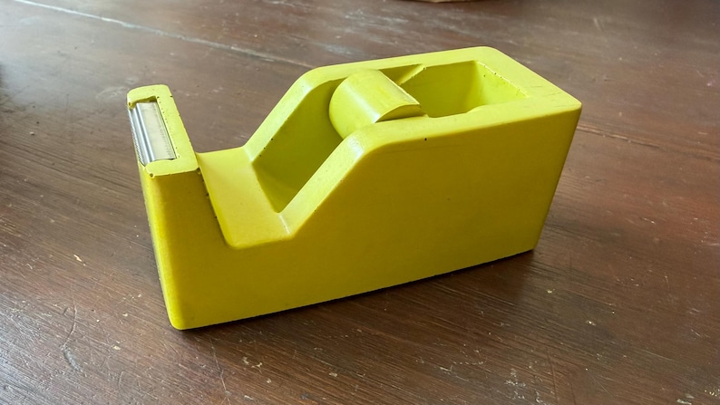 May include: A lime green tape dispenser featuring a smooth, solid-colored plastic body with a built-in metal serrated cutting blade at the front and a central cylindrical core designed to hold a standard roll of adhesive tape for office use.