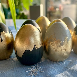 Könnte beinhalten: Eine Gruppe von acht dekorativen Eiern, die in Schwarz und Gold mit einem Metallic-Finish bemalt sind. Die Eier sind in einer Reihe auf einer blau-weißen Tischdecke angeordnet.