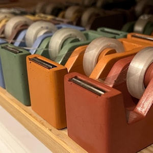 May include: A collection of rectangular tape dispensers in various matte colors including terracotta, orange, sage green, blue, and brown. Each dispenser features a clear roll of adhesive tape and a metal cutting blade at the front edge.