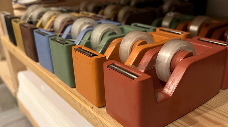 May include: A collection of rectangular tape dispensers in various matte colors including terracotta, orange, sage green, blue, and brown. Each dispenser features a clear roll of adhesive tape and a metal cutting blade at the front edge.