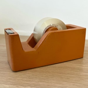 May include: A terracotta-colored tape dispenser with a matte, textured finish. The rectangular base holds a roll of clear adhesive tape, which is positioned on a central spindle. A small piece of tape is pulled forward to the metal cutting edge.
