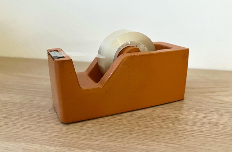 May include: A terracotta-colored tape dispenser with a matte, textured finish. The rectangular base holds a roll of clear adhesive tape, which is positioned on a central spindle. A small piece of tape is pulled forward to the metal cutting edge.