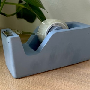 May include: A matte periwinkle blue desktop tape dispenser with a rectangular base and a slanted top profile. The dispenser holds a roll of clear adhesive tape, which is threaded through a serrated metal cutting blade at the front edge.