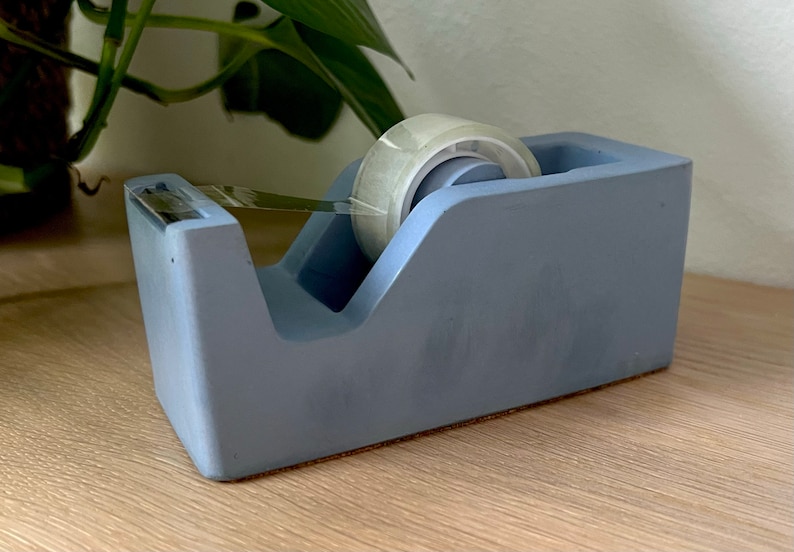 May include: A matte periwinkle blue desktop tape dispenser with a rectangular base and a slanted top profile. The dispenser holds a roll of clear adhesive tape, which is threaded through a serrated metal cutting blade at the front edge.