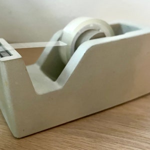 May include: A light gray desktop tape dispenser with a rectangular base and a slanted front edge. The dispenser holds a roll of clear adhesive tape, with a small portion of the tape pulled forward and resting across the serrated metal cutting blade.