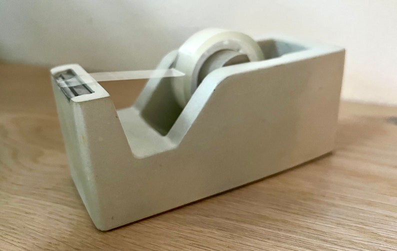 May include: A light gray desktop tape dispenser with a rectangular base and a slanted front edge. The dispenser holds a roll of clear adhesive tape, with a small portion of the tape pulled forward and resting across the serrated metal cutting blade.