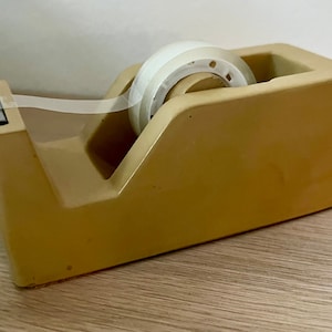 May include: A mustard yellow desktop tape dispenser with a smooth, matte finish. The dispenser holds a roll of clear adhesive tape, which extends forward to a metal serrated cutting blade positioned at the front edge of the plastic housing.