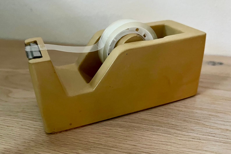 May include: A mustard yellow desktop tape dispenser with a smooth, matte finish. The dispenser holds a roll of clear adhesive tape, which extends forward to a metal serrated cutting blade positioned at the front edge of the plastic housing.