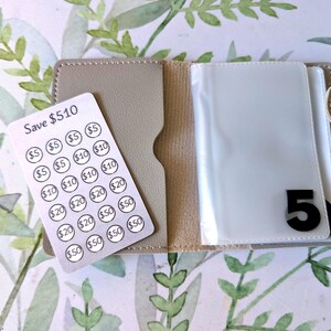 Può includere: Un portafoglio beige con chiusura a scatto, contenente una scheda di monitoraggio dei risparmi e custodie di plastica trasparente. La scheda di monitoraggio dei risparmi recita "Save $510" e presenta cerchi con importi in dollari. Il numero "5" è visibile sulla custodia.