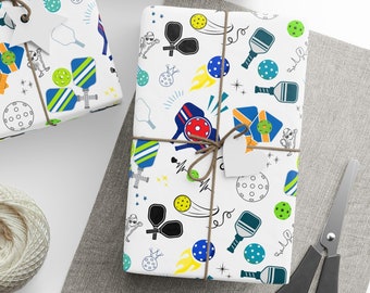 May include: White gift wrap with a repeating pattern of blue, green, and yellow pickleball paddles, balls, and other designs. The wrap is tied with brown twine and has a white gift tag.