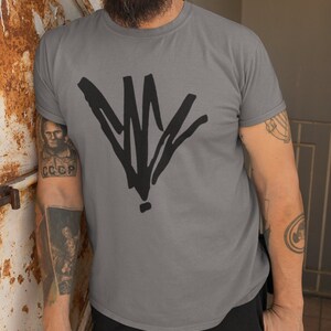 May include: Gray t-shirt with a large black abstract graphic on the front. The person in the image is wearing a blue baseball cap and black pants. The person has tattoos on their arms.