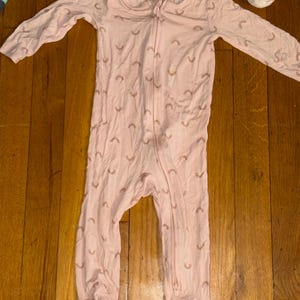 May include: A light pink baby sleeper with a full-length zipper and long sleeves. The sleeper is patterned with small, light brown rainbows. The footed sleeper is made of a soft material.