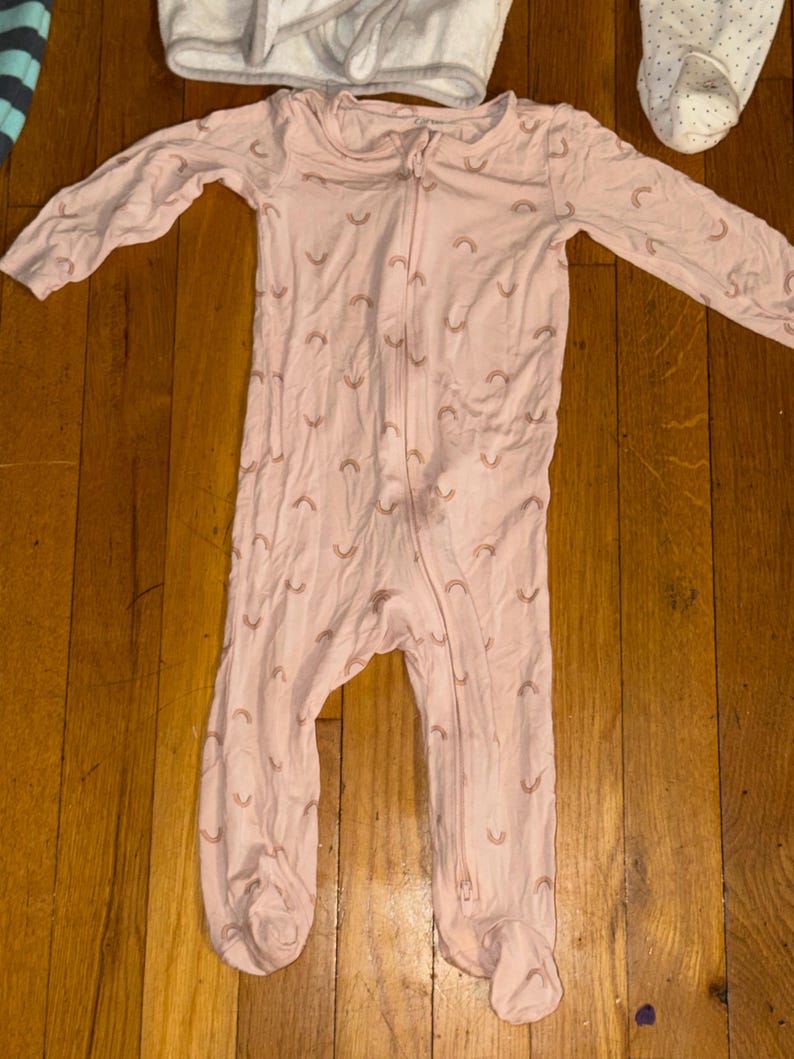 May include: A light pink baby sleeper with a full-length zipper and long sleeves. The sleeper is patterned with small, light brown rainbows. The footed sleeper is made of a soft material.