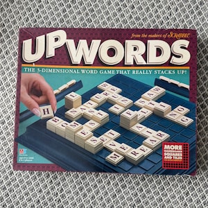 May include: A board game box for "Upwords" from the makers of Scrabble. The box is burgundy with the game title in large, cream-colored letters. The game board is dark blue, with beige letter tiles. The game is advertised as a 3-dimensional word game.