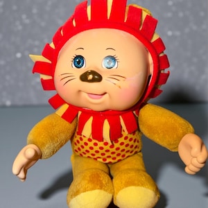 May include: A vintage plush lion doll with a red and yellow mane, a red and yellow spotted torso, and yellow limbs. The doll has blue eyes, a brown nose, and painted whiskers. The doll's arms are outstretched, and it has a smiling expression.