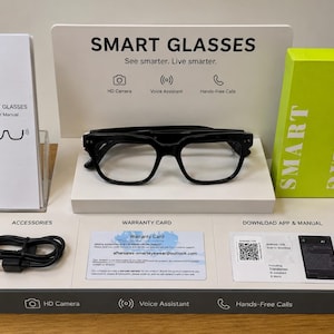 May include: Black smart glasses with a rectangular frame are displayed on a white platform. The display includes a user manual, a warranty card, a charging cable, and a box. The text on the display reads "SMART GLASSES" and "See smarter. Live smarter."