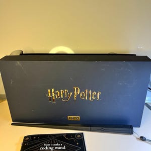 Puede incluir: Una caja azul oscuro con letras doradas que dicen "Harry Potter" y la marca "KANO". Una varita negra y un folleto titulado "How to make a coding wand" están frente a la caja. El folleto tiene diagramas y texto.