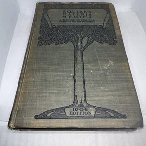 Puede incluir: Un libro antiguo titulado "Squibb's Materia Medica" con una ilustración dibujada a mano de un árbol en la portada. El libro es un manual para el médico y el farmacéutico y está fechado en 1906.