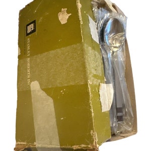 May include: A vintage olive green box of Stanley Roberts Inc. silverware, partially open, revealing wrapped stainless steel utensils. The box has a logo and text, with signs of wear and tear.