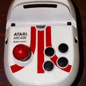 May include: White Atari Arcade Duo-Powered controller with a red joystick and red and white detailing. The controller features four black buttons and the Atari logo in red.