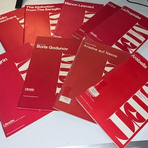 May include: A collection of red opera librettos, including titles like "Un Ballo in Maschera," "Boris Godunov," and "Arabella." The covers feature white text and the Metropolitan Opera logo. The books are scattered on a white surface.