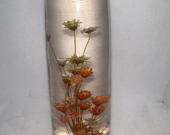 May include: A clear glass cylinder with dried flowers inside. The flowers are orange, white, and green. The cylinder is sitting on a wooden base with a gold label that says "Handcrafted".