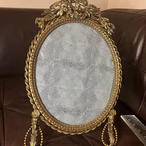 May include: An antique oval picture frame with a gold finish. The frame features a decorative bird and floral design at the top, with ornate detailing around the oval opening. The frame has a textured, light-colored backing.
