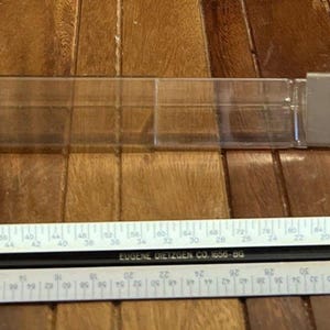 Puede incluir: Una caja de plástico transparente que contiene un triángulo de plástico Dietzgen y una regla blanca y negra con medidas en centímetros. La regla tiene el texto "Made in Germany" y "EAGLE DESIGNER CO. 600-30".