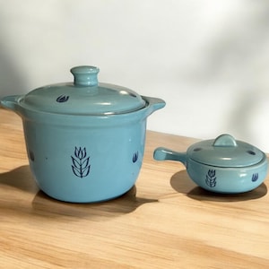 Può includere: Un set di due pezzi di pentolame in ceramica blu con un design a forma di grano. Il pezzo più grande è una pentola con coperchio, e il pezzo più piccolo è una casseruola coperta con manico.