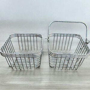 Puede incluir: Dos cestas de alambre plateado, una con asa, sobre una superficie de madera clara. Las cestas son cuadradas y tienen un patrón de rejilla.