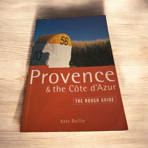 May include: A red, orange, and white book titled "Provence & the Côte d'Azur" with the words "The Rough Guide" in a white box. The cover features a photo of a white and yellow road sign with the numbers "56" and "10" on it.