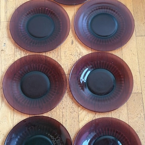 Puede incluir: Un juego de seis platos de vidrio vintage en color ciruela oscuro. Cada plato presenta un centro oscuro y un diseño texturizado y radiante alrededor del borde. Los platos están dispuestos sobre una superficie de madera.