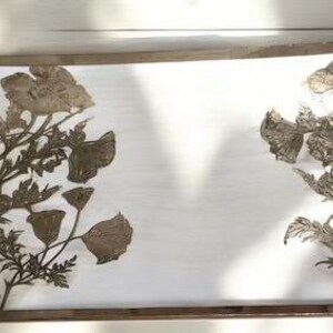 Könnte beinhalten: Ein rechteckiges Serviertablett mit weißem Hintergrund und goldfarbenem Metallrand. Das Tablett zeigt zwei Blumendesigns mit in die Oberfläche gepresstem Blattgold.