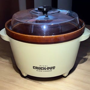 May include: A Rival Crock-Pot slow cooker with a cream-colored exterior, brown rim, and clear lid. The slow cooker has a black handle on the lid and the words "RIVAL CROCK-POT SLOW-COOKER" printed on the front.