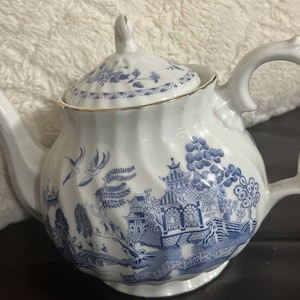 Puede incluir: Tetera de porcelana blanca con diseño azul de sauce. La tetera presenta un asa curva, un pico con ribete dorado y una tapa con diseño floral. El cuerpo de la tetera tiene una textura acanalada.