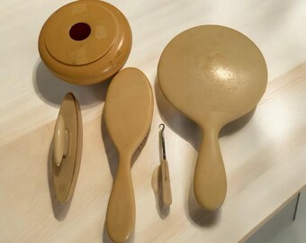 Può includere: Un set di cinque articoli da toeletta vintage di colore beige chiaro. Il set comprende una cipria rotonda, uno specchio da mano, una spazzola per capelli, un piccolo oggetto di forma ovale e un piccolo gancio di metallo.