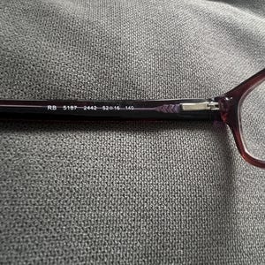 May include: Close-up of a pair of eyeglasses with a dark burgundy frame. The text "RB 5187 2442 52▢16 140" is printed on the temple. The glasses are set against a gray textured background.