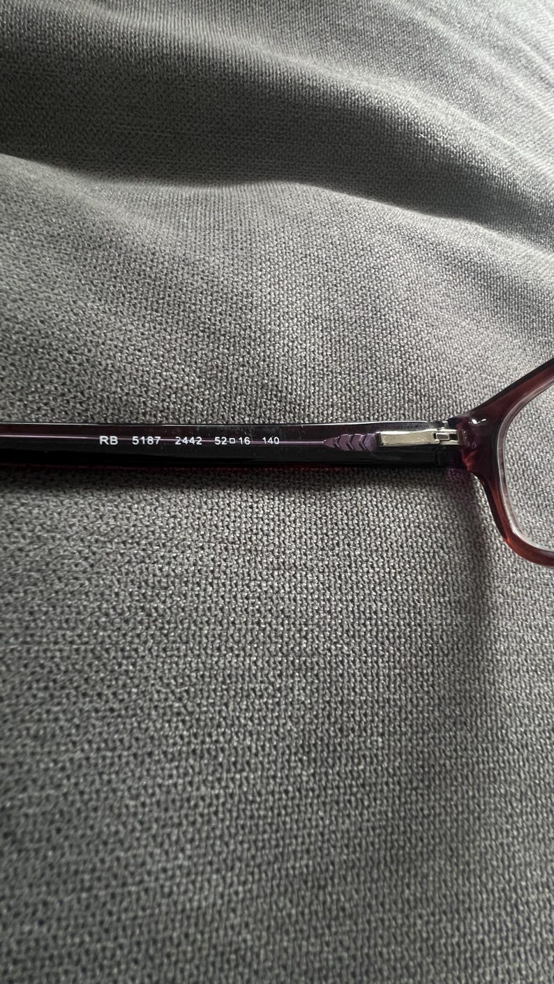 May include: Close-up of a pair of eyeglasses with a dark burgundy frame. The text "RB 5187 2442 52▢16 140" is printed on the temple. The glasses are set against a gray textured background.