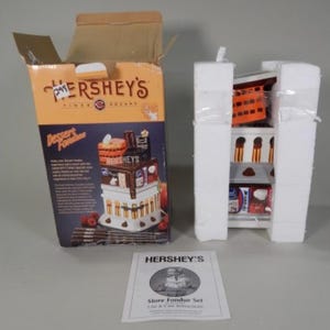 May include: A Hershey's Times Square Dessert Fondue set, packaged in a cardboard box. The box features a colorful illustration of the fondue set, with the Hershey's logo prominently displayed. The set includes a ceramic fondue pot and accessories.