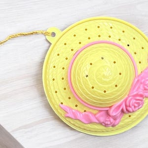 Puede incluir: Un sombrero de plástico amarillo con una cinta rosa y adornos de flores. El sombrero tiene un patrón de agujeros y una cuerda para colgar.