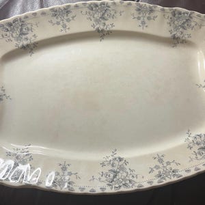 May include: A rectangular, off-white serving platter with a scalloped edge. The platter is decorated with a delicate floral pattern in shades of gray. The floral design is concentrated at the corners and along the rim, creating an elegant aesthetic.