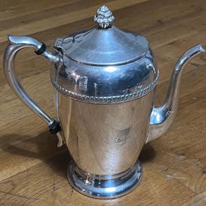 Puede incluir: Una tetera de plata con un pomo decorativo en la tapa y asas negras. La tetera tiene un cuerpo redondo y una base acampanada.